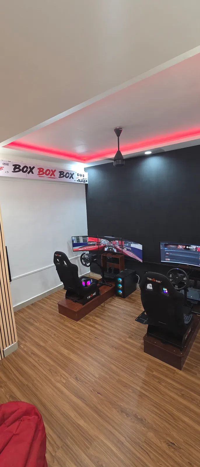 The floor at BoxBoxBox Guindy — both racing simulator rigs side by side under a BOX BOX BOX banner and a red LED ceiling cove, with a beanbag on the wooden floor for whoever is waiting.