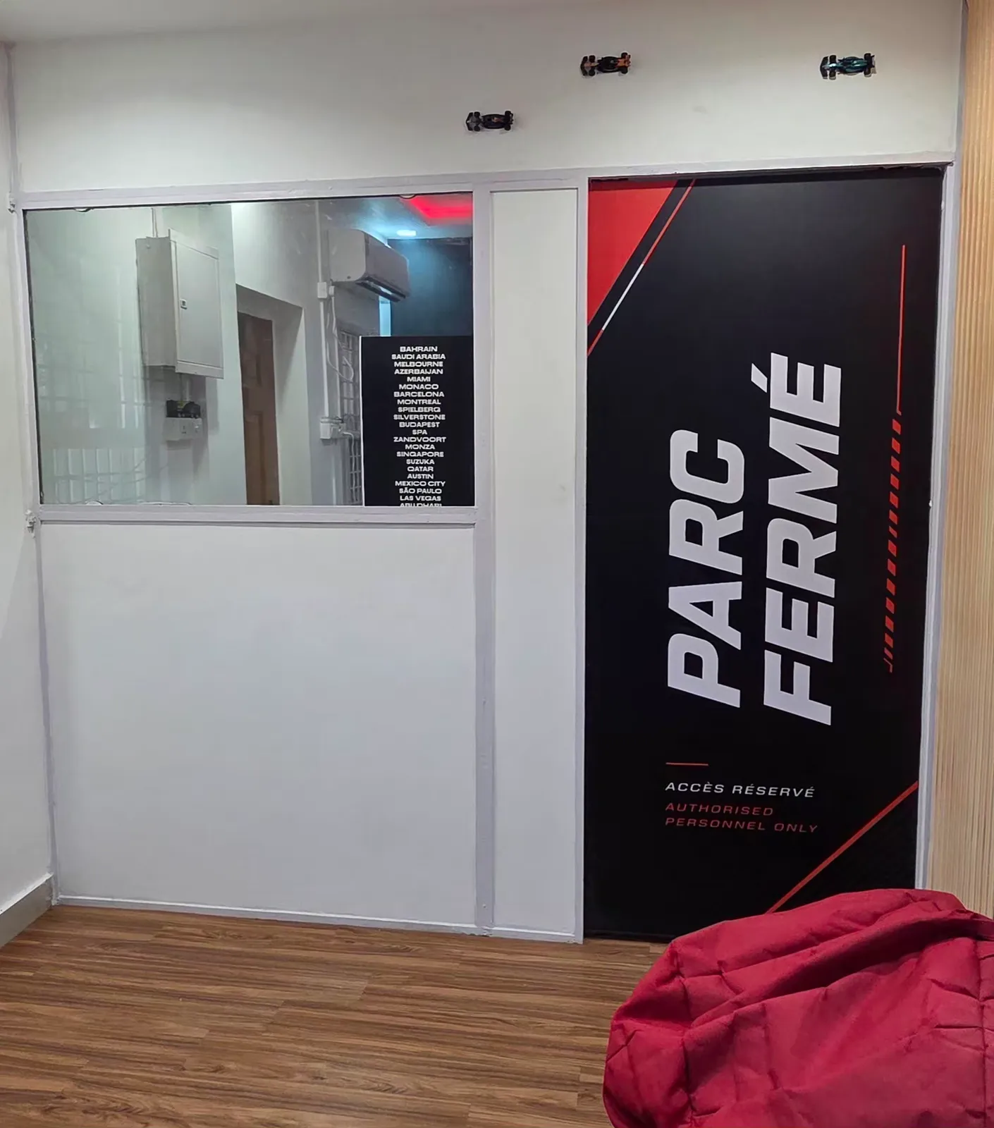 The waiting area at BoxBoxBox Guindy — a PARC FERMÉ wall graphic beside a window into the back room, model F1 cars along the wall, a board listing the season's circuits, and a red beanbag.
